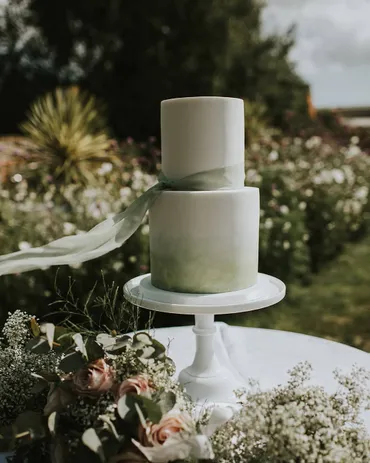 Two-tier sage green ombre wedding cake with silk ribbon and blush roses on a white stand in a garden